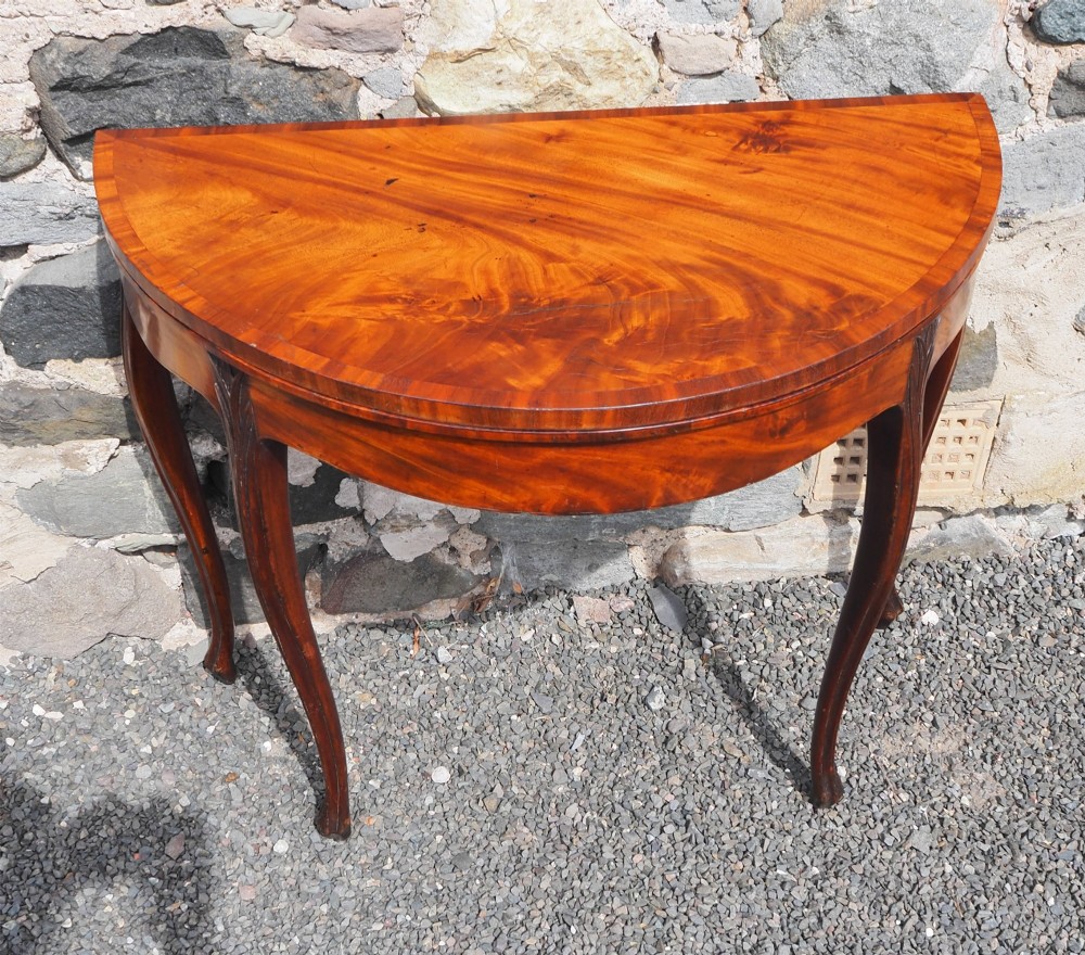 An 18th-century demilune folding card table crafted with flame mahogany veneer and carved slender legs.