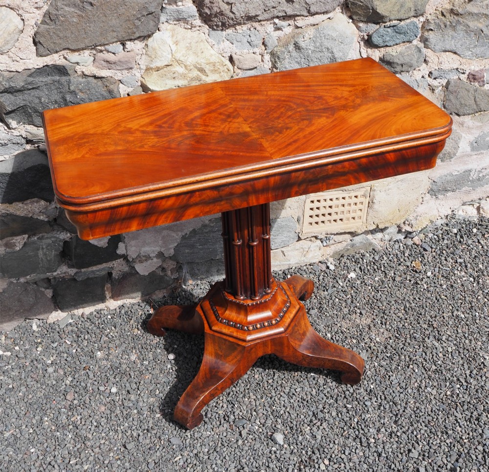 An 1830s William IV flame mahogany tea table featuring a fold-over rectangular top set on a fluted pedestal column with a four-point platform base and scroll feet.