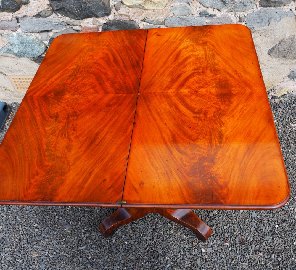 An 1830s William IV flame mahogany tea table featuring a fold-over rectangular top set on a fluted pedestal column with a four-point platform base and scroll feet. View X