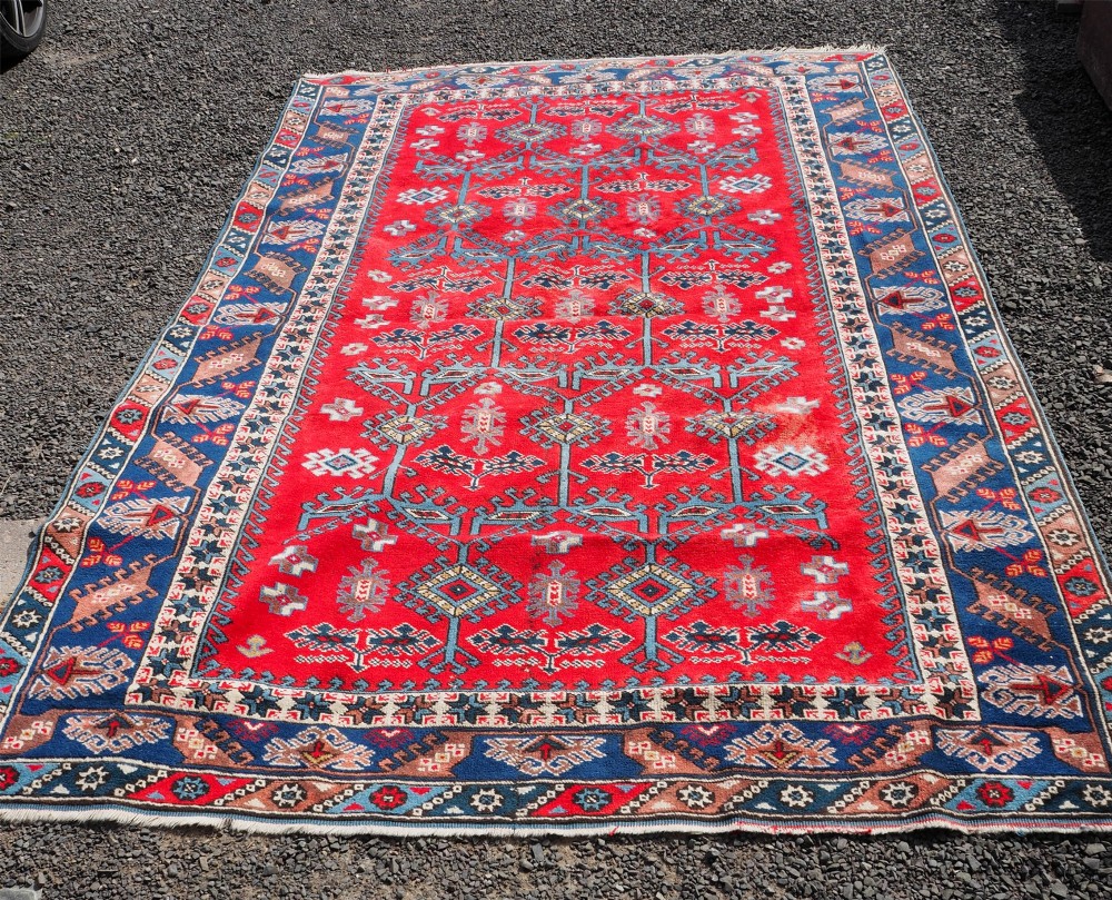A large rectangular hand-knotted wool carpet featuring a crimson red field with geometric blue and off-white motifs framed by a wide navy blue decorative border. View II