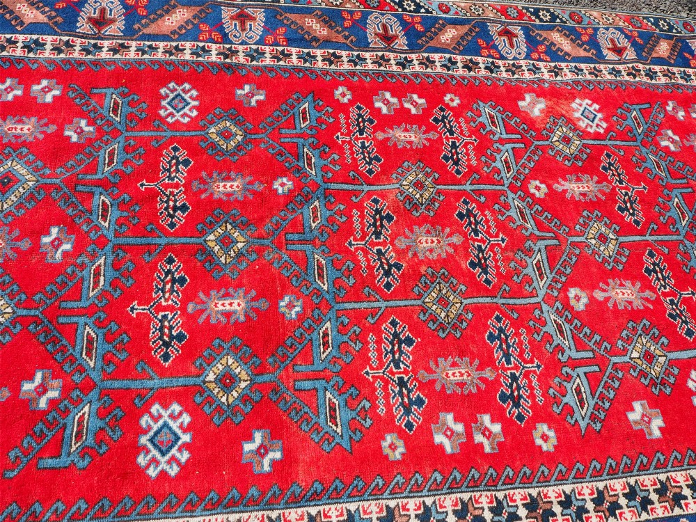 A large rectangular hand-knotted wool carpet featuring a crimson red field with geometric blue and off-white motifs framed by a wide navy blue decorative border. View V