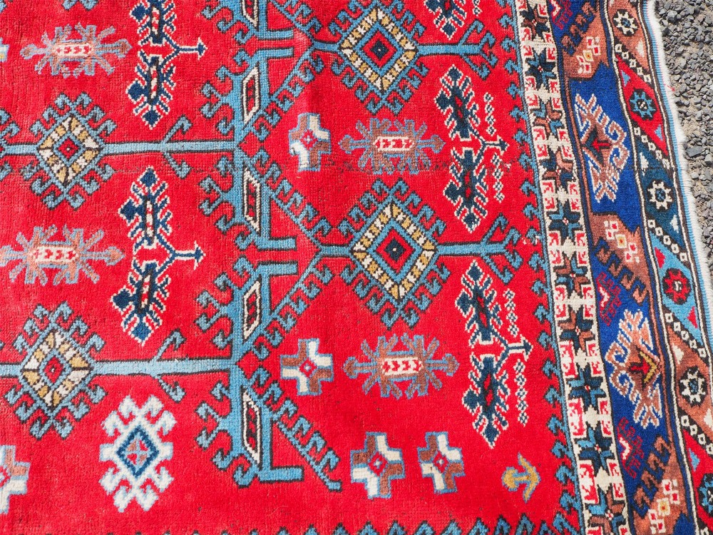 A large rectangular hand-knotted wool carpet featuring a crimson red field with geometric blue and off-white motifs framed by a wide navy blue decorative border. View VI