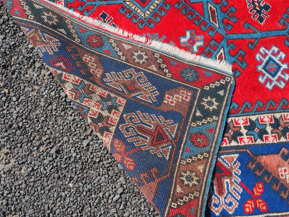A large rectangular hand-knotted wool carpet featuring a crimson red field with geometric blue and off-white motifs framed by a wide navy blue decorative border. View VII