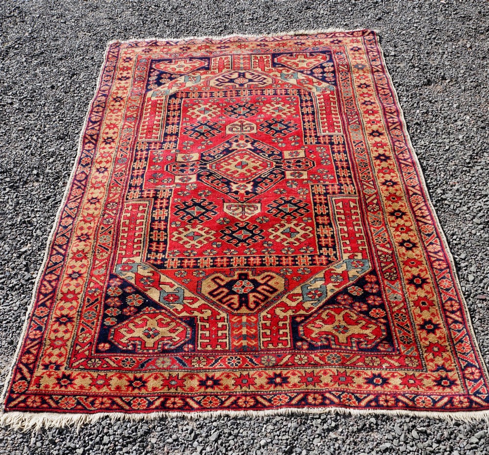 An antique hand-knotted Persian wool rug featuring a central geometric diamond medallion in navy blue set against a deep red field with patterned borders. View II