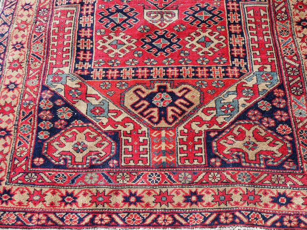 An antique hand-knotted Persian wool rug featuring a central geometric diamond medallion in navy blue set against a deep red field with patterned borders. View V