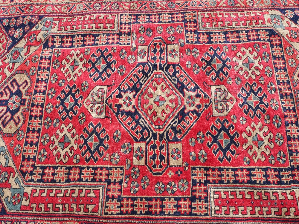 An antique hand-knotted Persian wool rug featuring a central geometric diamond medallion in navy blue set against a deep red field with patterned borders. View VI