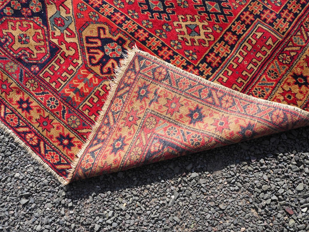 An antique hand-knotted Persian wool rug featuring a central geometric diamond medallion in navy blue set against a deep red field with patterned borders. View VII