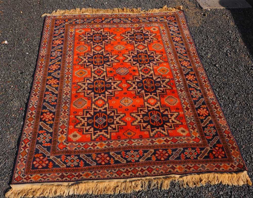 A rectangular hand-knotted wool rug featuring a bright terracotta field with dark navy geometric star medallions and detailed outer borders.