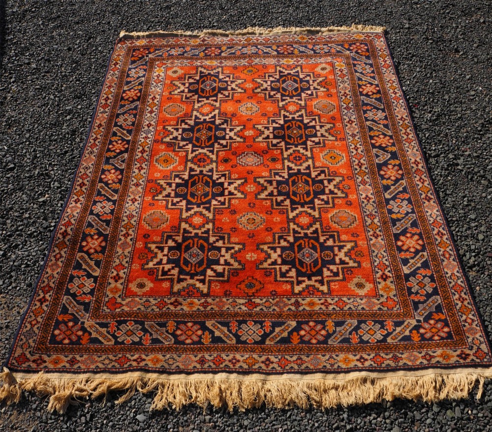 A rectangular hand-knotted wool rug featuring a bright terracotta field with dark navy geometric star medallions and detailed outer borders. View II