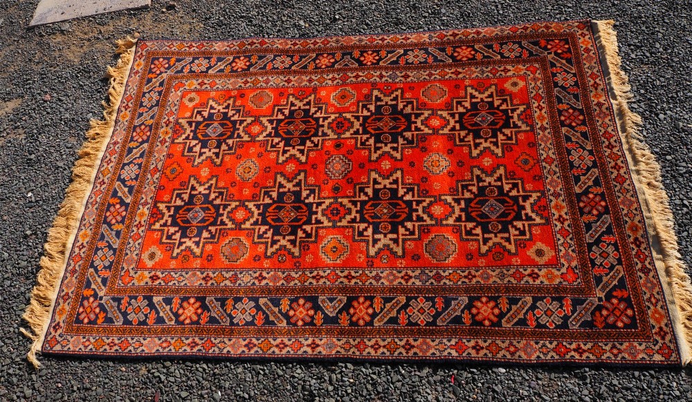 A rectangular hand-knotted wool rug featuring a bright terracotta field with dark navy geometric star medallions and detailed outer borders. View III