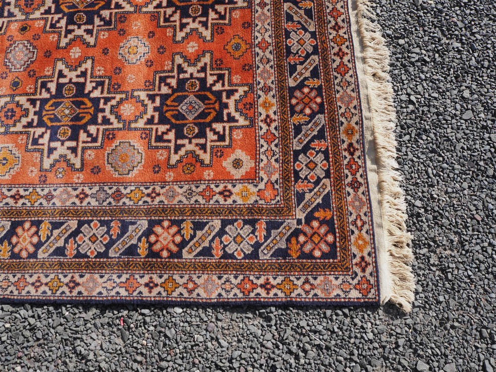 A rectangular hand-knotted wool rug featuring a bright terracotta field with dark navy geometric star medallions and detailed outer borders. View IV