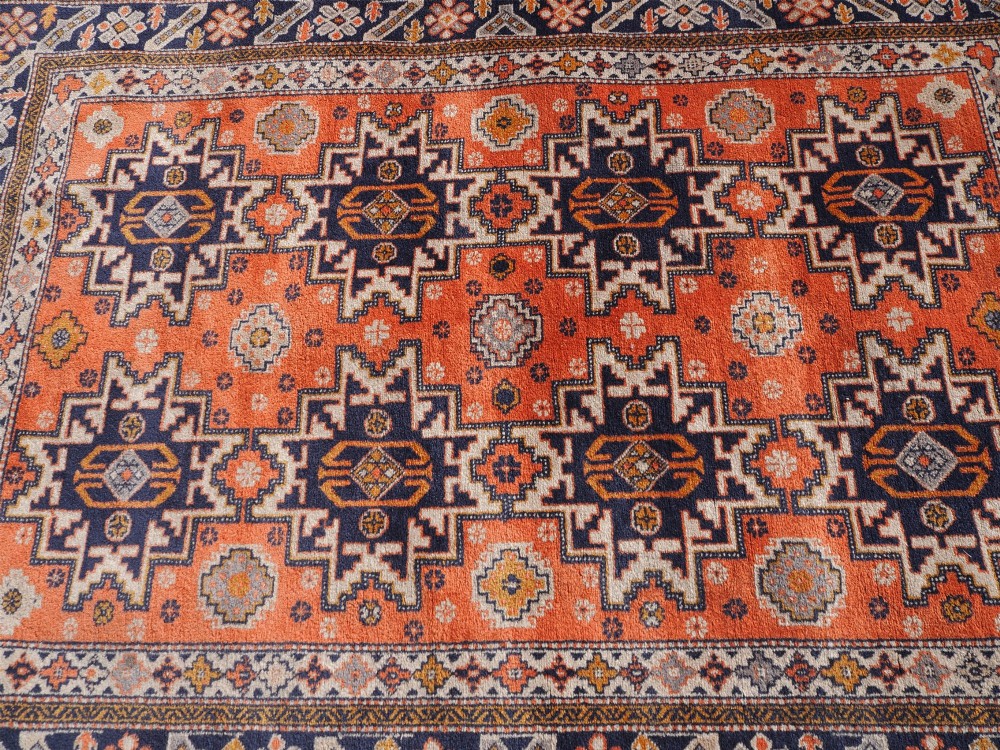 A rectangular hand-knotted wool rug featuring a bright terracotta field with dark navy geometric star medallions and detailed outer borders. View V