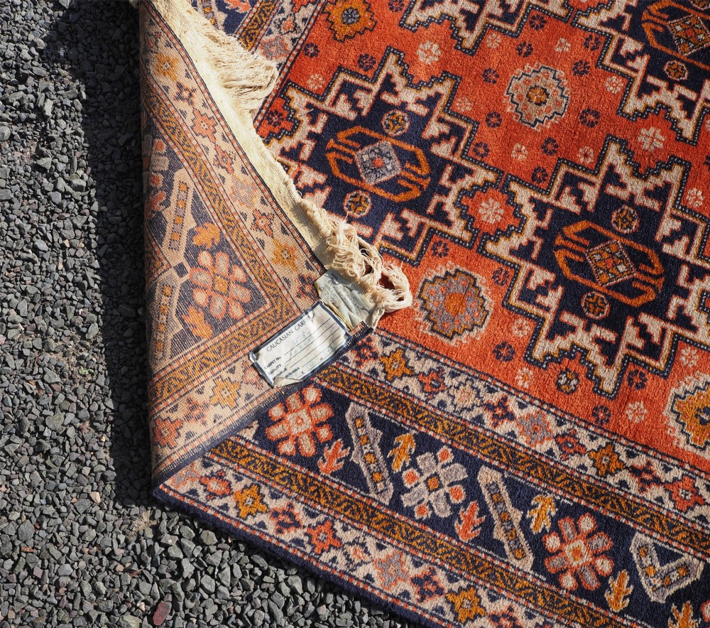 A rectangular hand-knotted wool rug featuring a bright terracotta field with dark navy geometric star medallions and detailed outer borders. View VI
