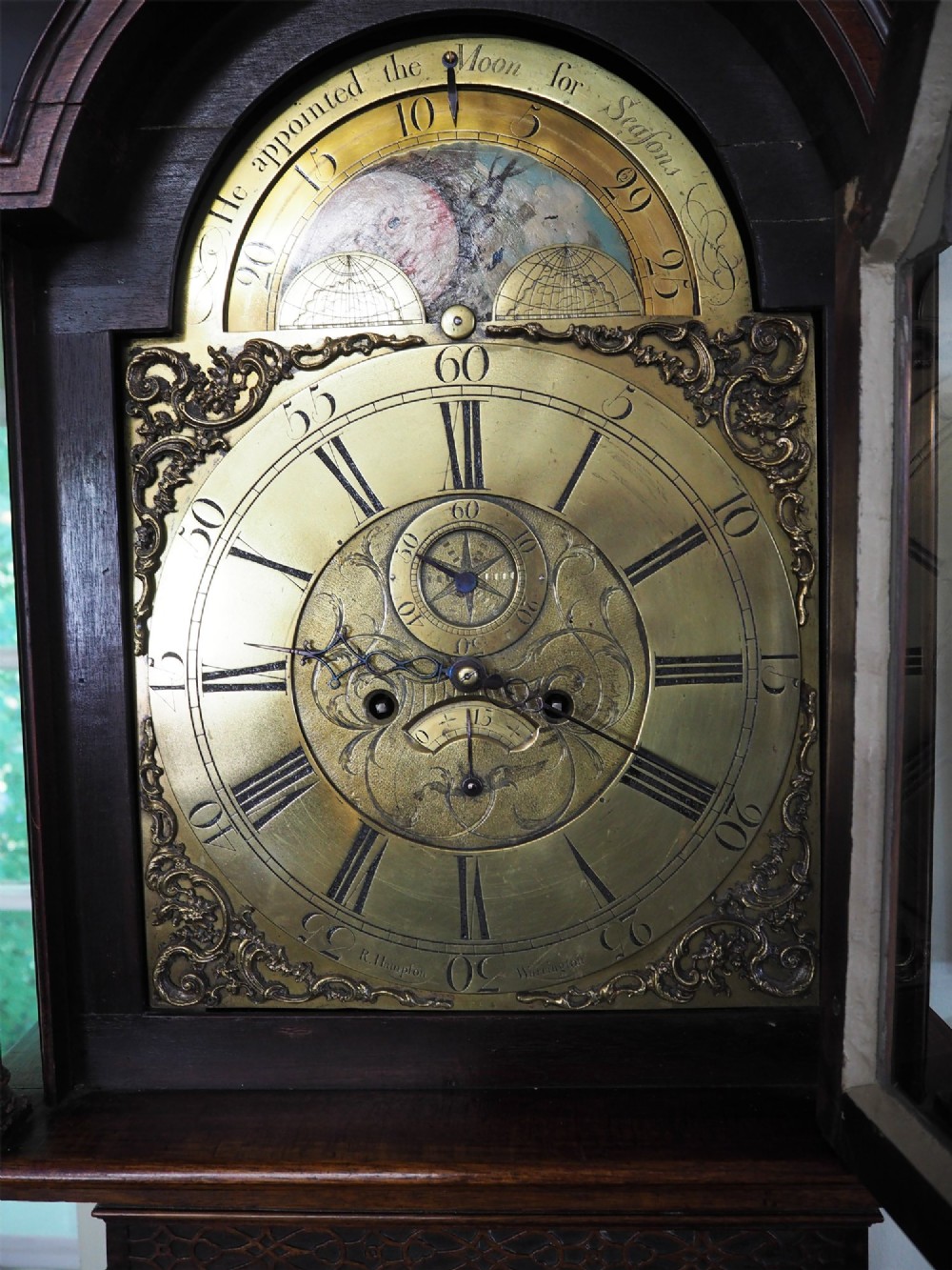 An eighteenth-century mahogany longcase clock featuring a swan-neck pediment with brass finials, an ornate brass dial with a moon phase arch, and a flame mahogany veneer trunk door. View VI
