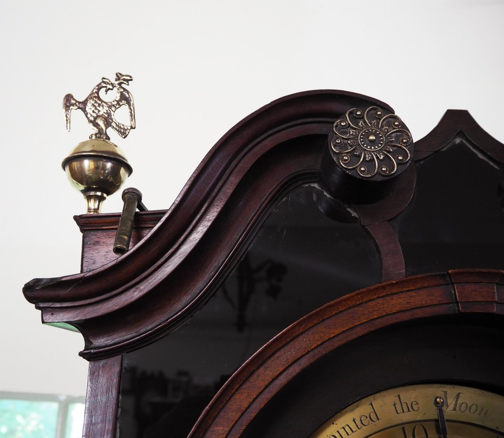 An eighteenth-century mahogany longcase clock featuring a swan-neck pediment with brass finials, an ornate brass dial with a moon phase arch, and a flame mahogany veneer trunk door. View VIII