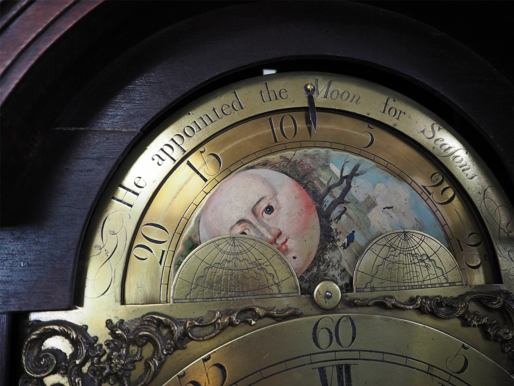 An eighteenth-century mahogany longcase clock featuring a swan-neck pediment with brass finials, an ornate brass dial with a moon phase arch, and a flame mahogany veneer trunk door. View IX