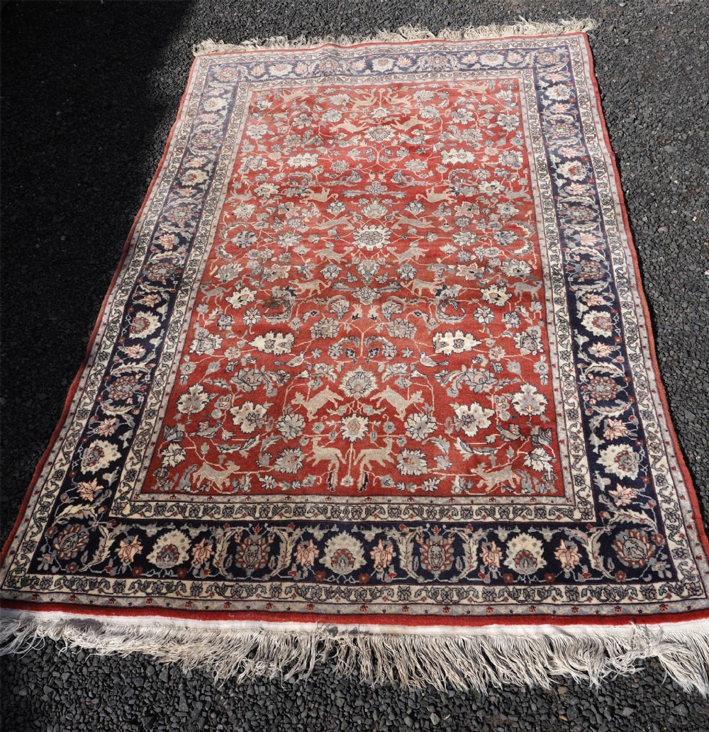 A rectangular hand-knotted wool rug featuring a rich rust red central field patterned with animal and floral motifs, framed by a dark navy blue ornate border with fringed edges. View II