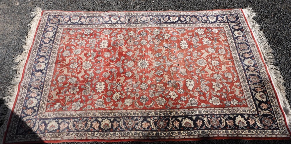 A rectangular hand-knotted wool rug featuring a rich rust red central field patterned with animal and floral motifs, framed by a dark navy blue ornate border with fringed edges. View III