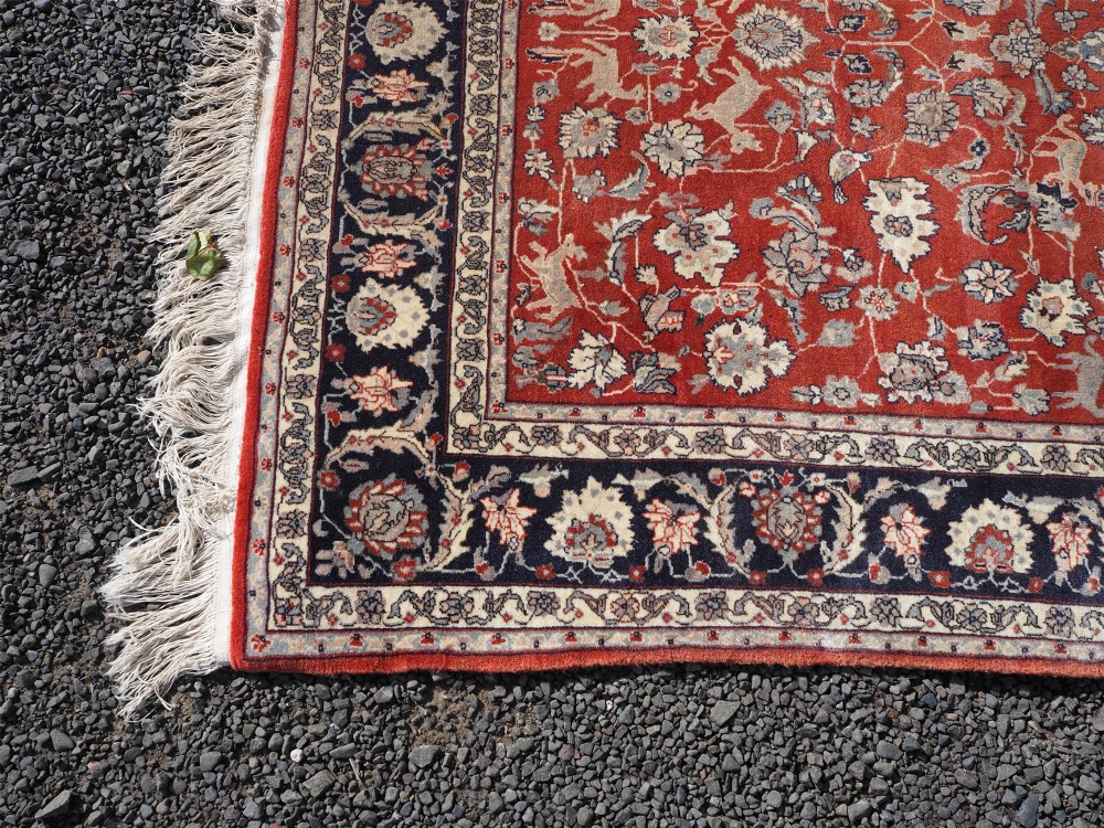 A rectangular hand-knotted wool rug featuring a rich rust red central field patterned with animal and floral motifs, framed by a dark navy blue ornate border with fringed edges. View IV