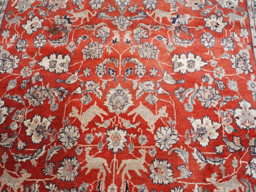A rectangular hand-knotted wool rug featuring a rich rust red central field patterned with animal and floral motifs, framed by a dark navy blue ornate border with fringed edges. View V