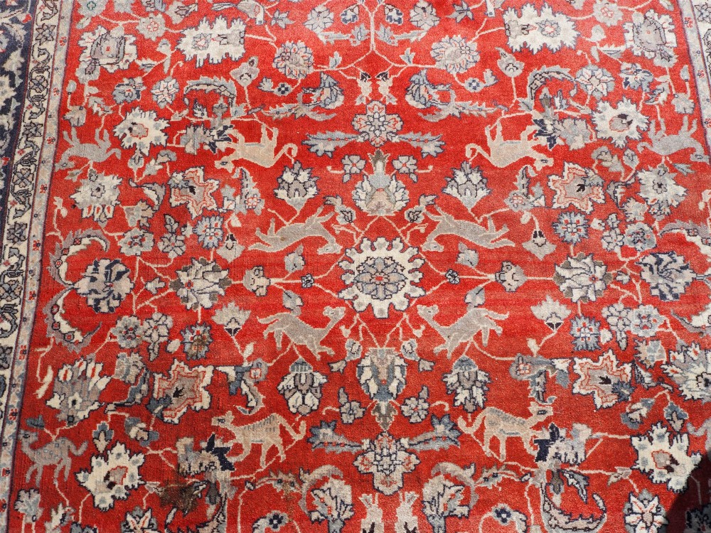 A rectangular hand-knotted wool rug featuring a rich rust red central field patterned with animal and floral motifs, framed by a dark navy blue ornate border with fringed edges. View VI