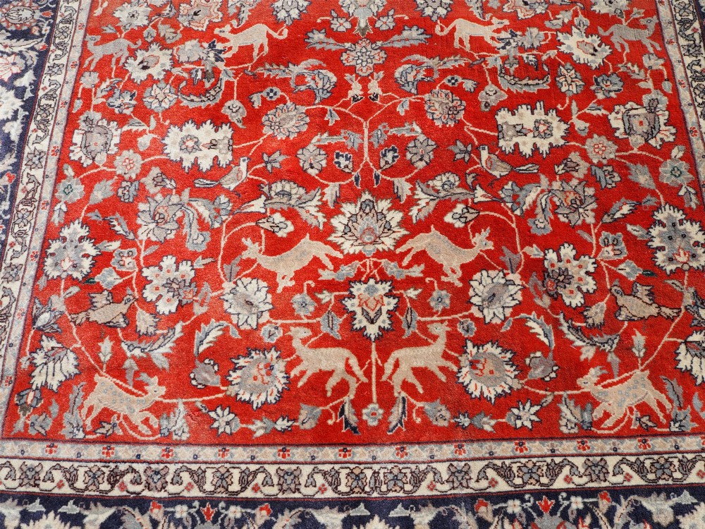 A rectangular hand-knotted wool rug featuring a rich rust red central field patterned with animal and floral motifs, framed by a dark navy blue ornate border with fringed edges. View VII