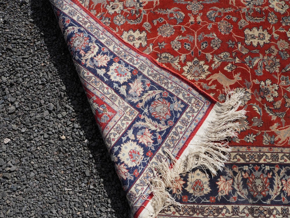 A rectangular hand-knotted wool rug featuring a rich rust red central field patterned with animal and floral motifs, framed by a dark navy blue ornate border with fringed edges. View VIII