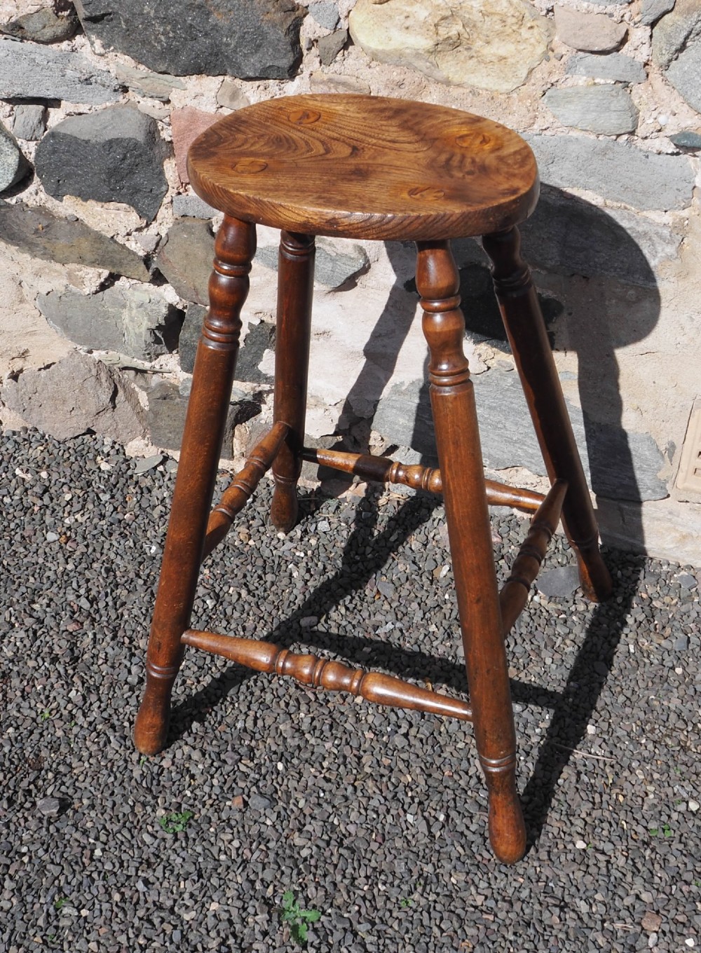 A 19th-century high stool constructed from ash and beech timber, featuring turned splayed legs, foot stretchers, and a rounded wooden seat.