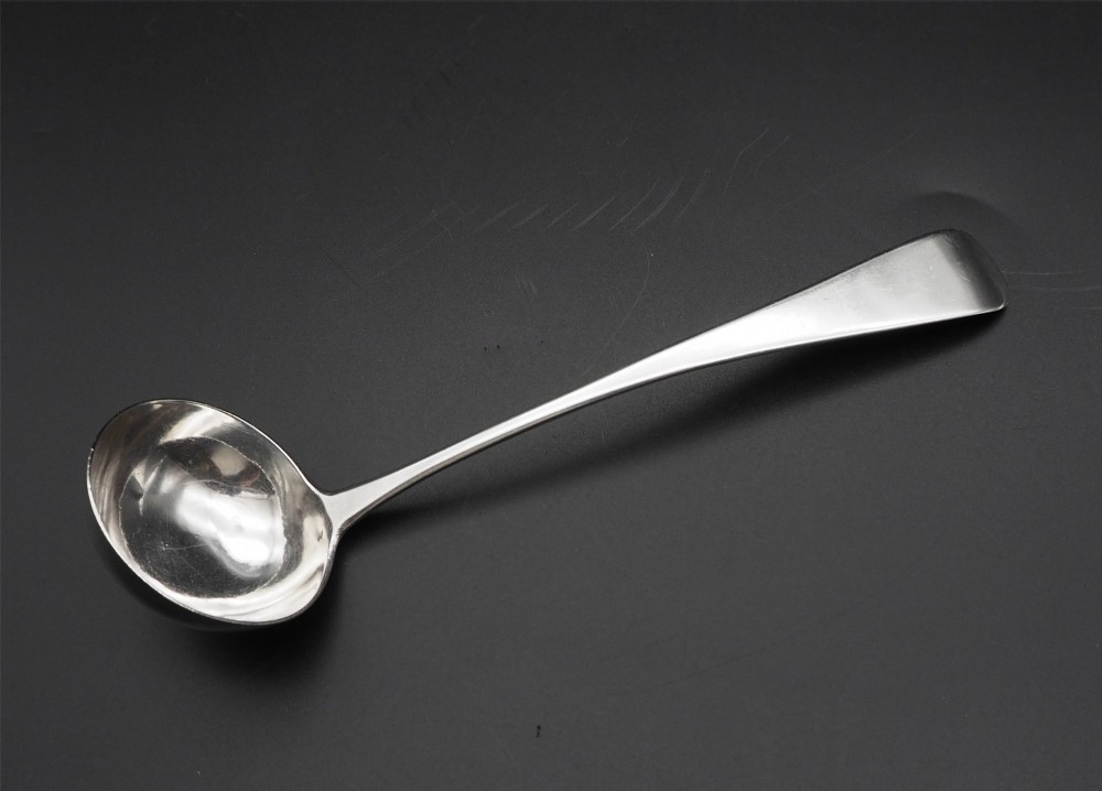 An antique silver sauce ladle with a deep circular bowl and a slender plain stem against a dark background.
