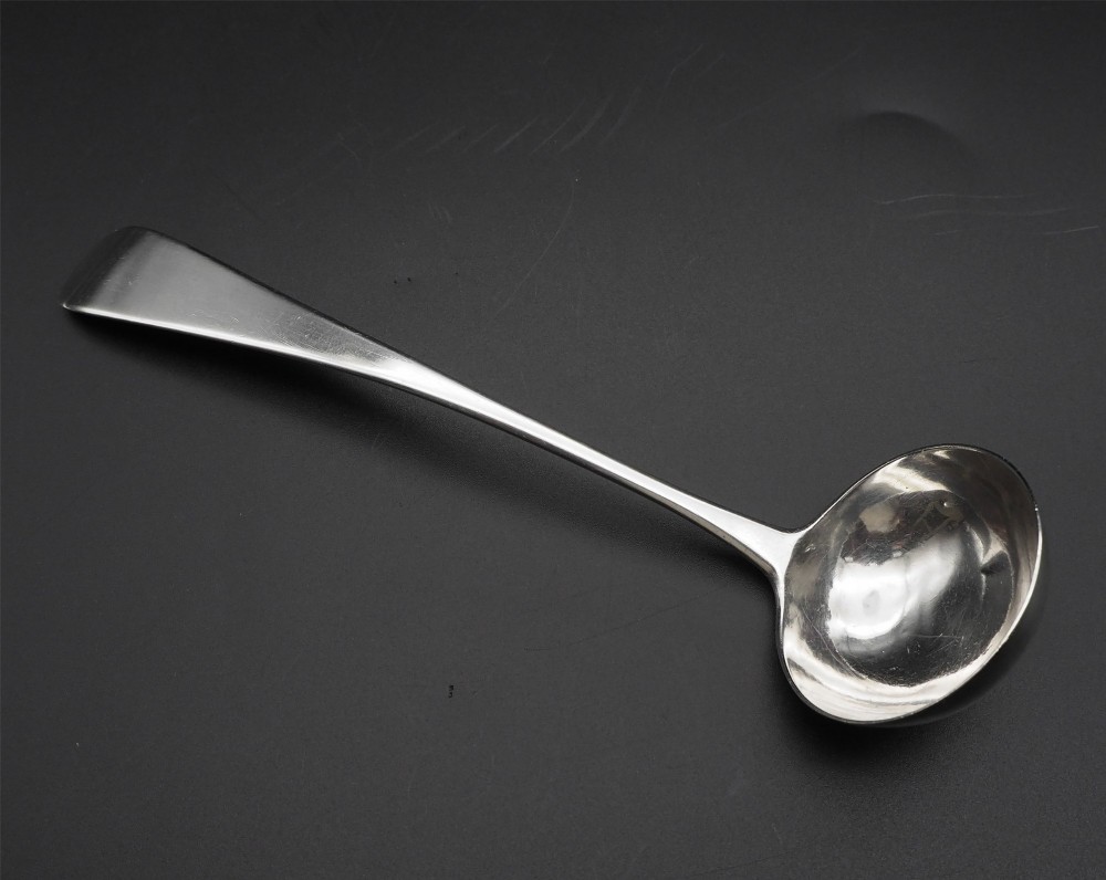 An antique silver sauce ladle with a deep circular bowl and a slender plain stem against a dark background. View II
