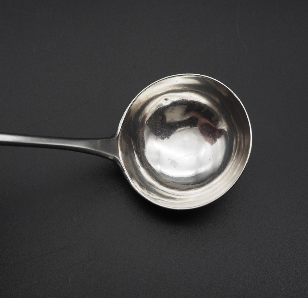 An antique silver sauce ladle with a deep circular bowl and a slender plain stem against a dark background. View III