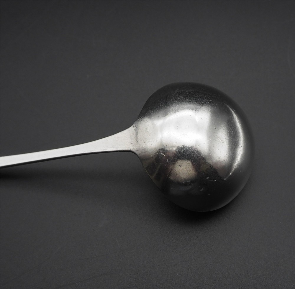 An antique silver sauce ladle with a deep circular bowl and a slender plain stem against a dark background. View IV