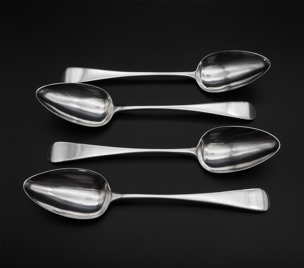 A set of four polished antique silver serving spoons with smooth handles and oval bowls arranged on a dark backdrop.