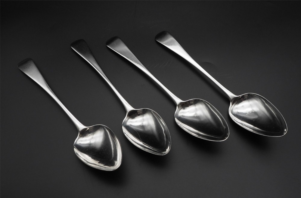 A set of four polished antique silver serving spoons with smooth handles and oval bowls arranged on a dark backdrop. View II