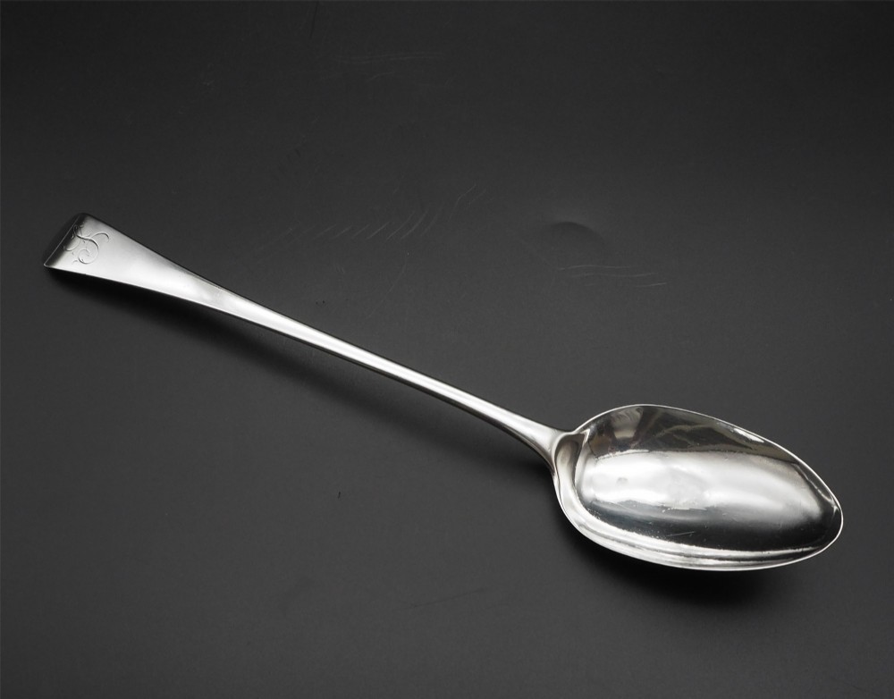 A Georgian sterling silver basting spoon featuring a long tapered handle with an engraved crest and a polished oval bowl.