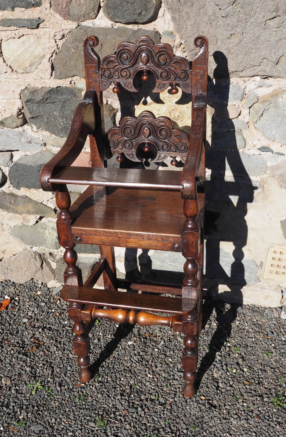 A dark oak childs high chair featuring double carved scroll cresting panels, turned legs, a footrest, and a front bar.