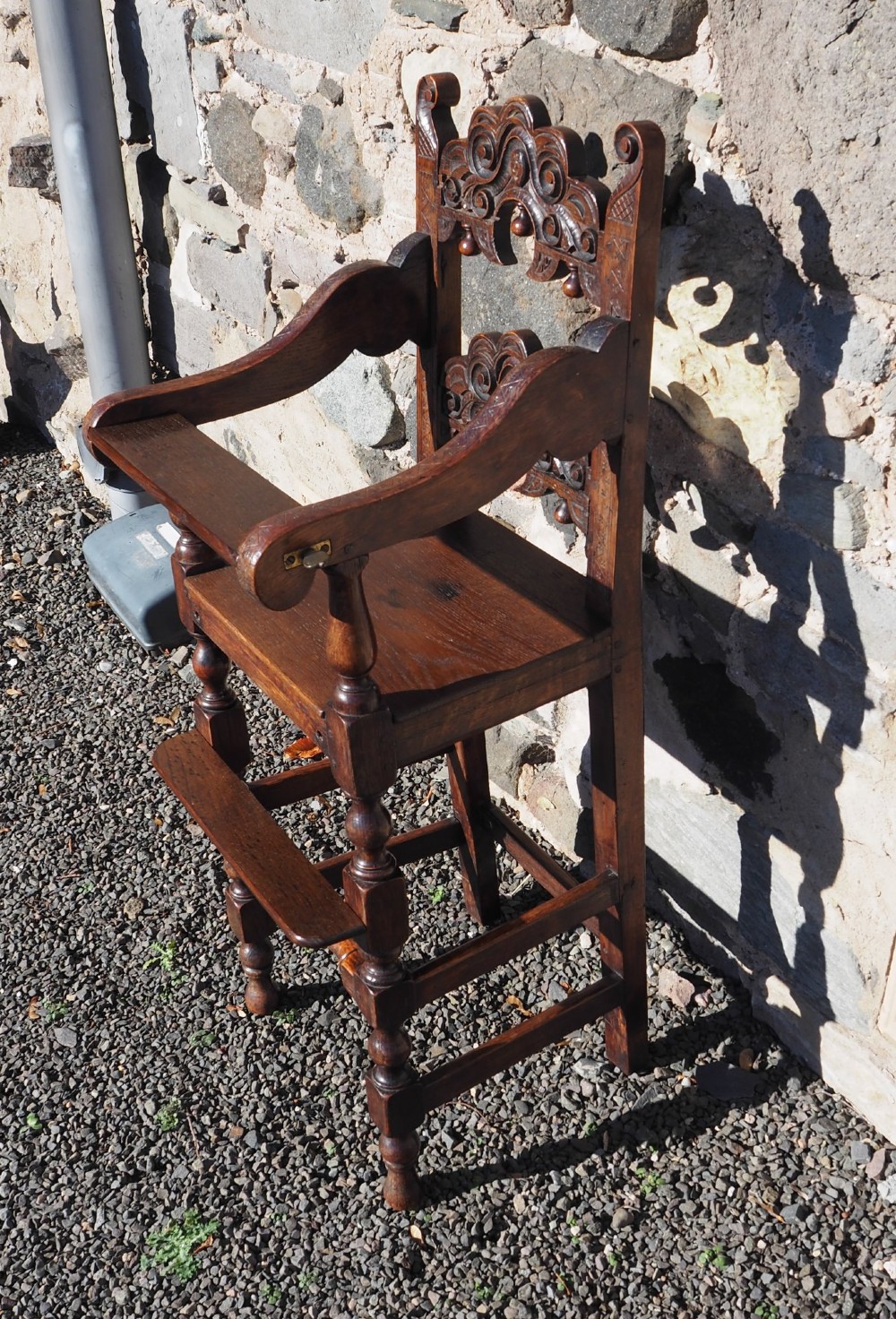 A dark oak childs high chair featuring double carved scroll cresting panels, turned legs, a footrest, and a front bar. View II