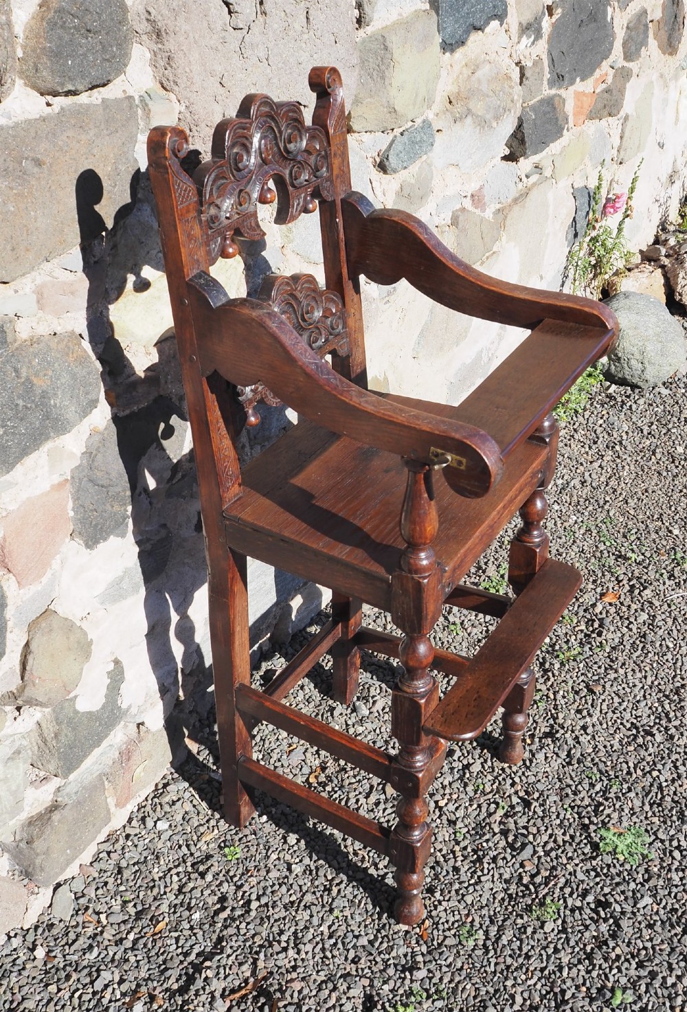 A dark oak childs high chair featuring double carved scroll cresting panels, turned legs, a footrest, and a front bar. View III
