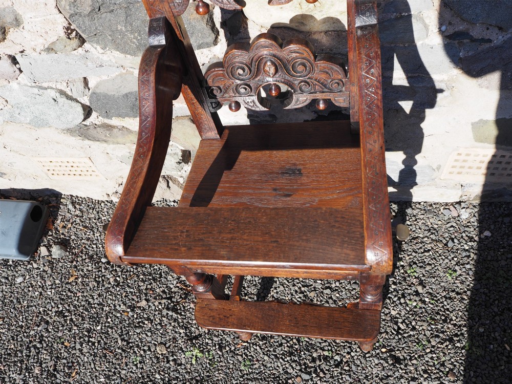 A dark oak childs high chair featuring double carved scroll cresting panels, turned legs, a footrest, and a front bar. View V
