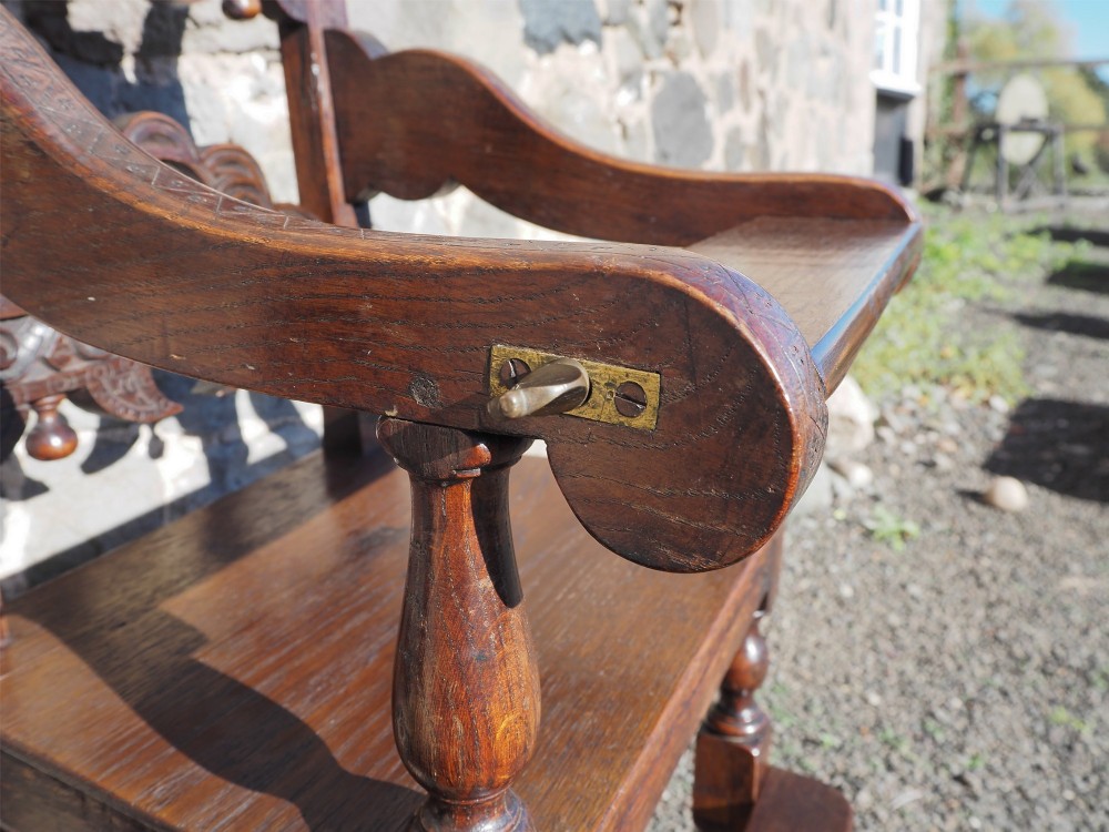 A dark oak childs high chair featuring double carved scroll cresting panels, turned legs, a footrest, and a front bar. View VII