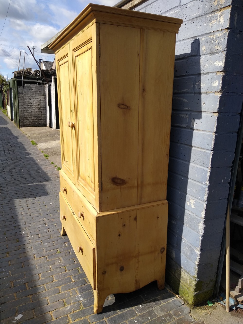 A tall natural pine linen press featuring a two-door upper cupboard above two short drawers and one long drawer with turned wooden knobs. View VII
