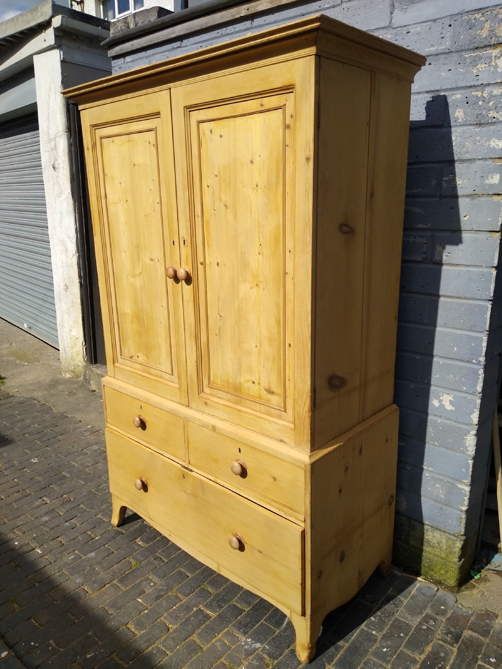 A tall natural pine linen press featuring a two-door upper cupboard above two short drawers and one long drawer with turned wooden knobs. View VIII