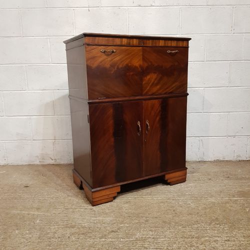 fig 1. An Art Deco mahogany drinks cabinet featuring bookmatched wood grain veneers, brass handles, a upper drop-front compartment, lower cupboard doors, and a stepped plinth base.