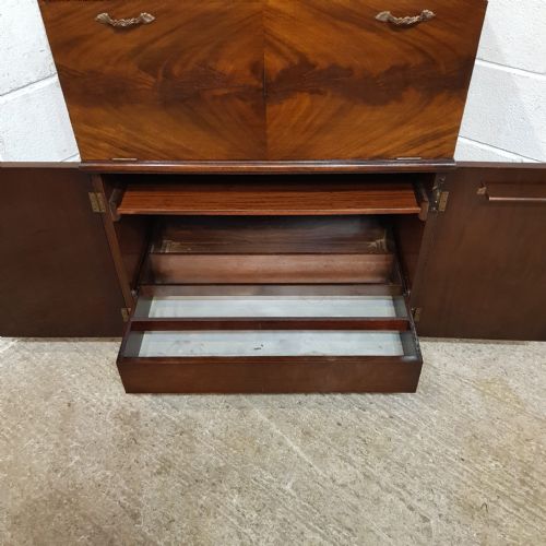 fig 2. An Art Deco mahogany drinks cabinet featuring bookmatched wood grain veneers, brass handles, a upper drop-front compartment, lower cupboard doors, and a stepped plinth base.