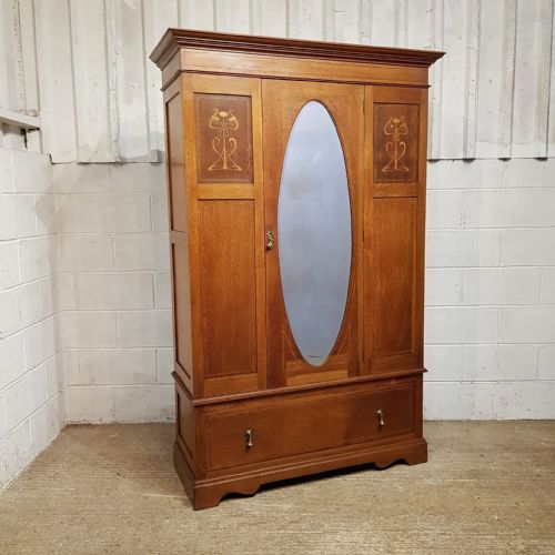 fig 1. An Arts and Crafts solid oak wardrobe featuring a central door with an oval glass mirror, flanking panels with inlaid botanical marquetry, and a single drawer at the base.