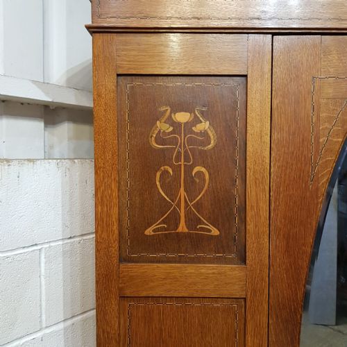fig 2. An Arts and Crafts solid oak wardrobe featuring a central door with an oval glass mirror, flanking panels with inlaid botanical marquetry, and a single drawer at the base.