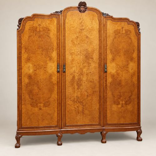 fig 1. An antique circa 1920 burr walnut triple wardrobe featuring three panelled doors with figured wood grain, carved top cresting, and carved cabriole feet.