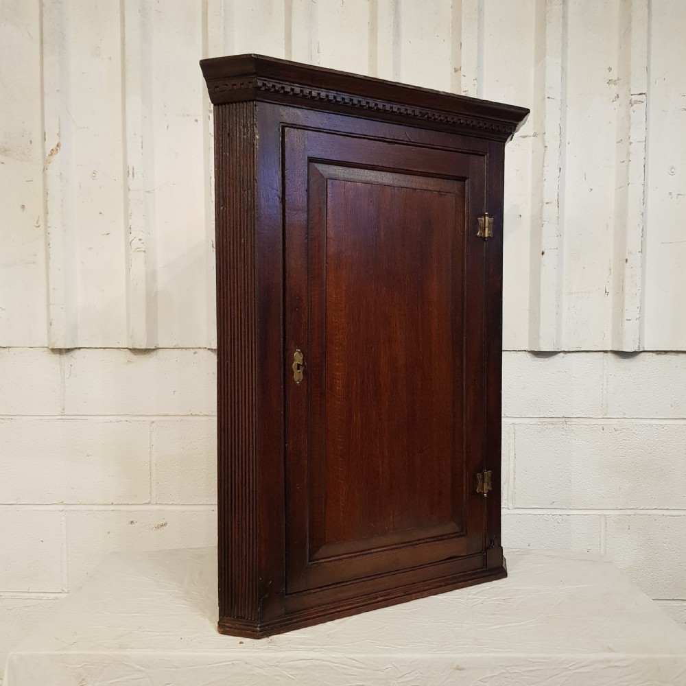 An 18th-century dark oak wall-hanging corner cabinet featuring a single panelled door with brass H-hinges and a keyhole escutcheon, topped with a dentil moulded cornice. View II