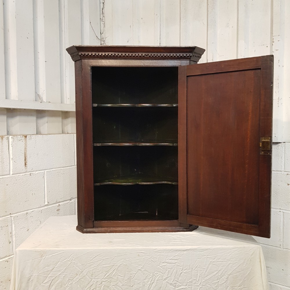 An 18th-century dark oak wall-hanging corner cabinet featuring a single panelled door with brass H-hinges and a keyhole escutcheon, topped with a dentil moulded cornice. View IV