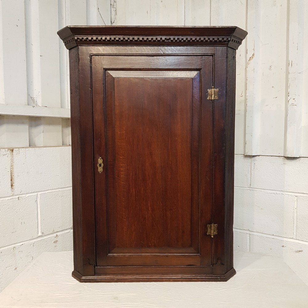 An 18th-century dark oak wall-hanging corner cabinet featuring a single panelled door with brass H-hinges and a keyhole escutcheon, topped with a dentil moulded cornice. View V